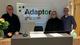 Foto av 3 ansatte fra AdaptorGruppen som står i resepsjonen foran et skilt med AdaptorGruppen sin logo.